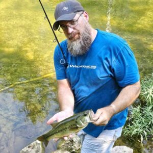 Bearded man in a blue Wilderness Systems shirt holding a small bass in a clear creek; identified as Andy Dill, known as The Bearded Paddler on YouTube.