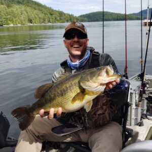Smiling kayak angler Tyler S. holding a large largemouth bass with fishing rods and a Ketch board visible on the kayak in the background.