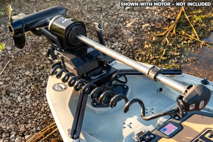 Close-up of a bow-mounted trolling motor plate installed on a kayak, with coiled wiring and mounting hardware visible. The motor is attached for demonstration but not included with the mount. The kayak is resting on rocky shoreline, with a 'Made in USA' sticker visible near the mount.